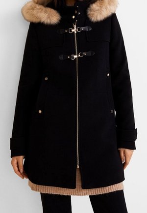 Czarny płaszcz średniej długości z beżowym sztucznym futrzanym kołnierzem, z przodu zamek błyskawiczny i dwa zapięcia na klamry, noszony na beżowym dzianinowym swetrze.