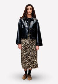 Vrouw staat gekleed in een zwart leren jack, een midirok met luipaardprint, zwarte sokken en een paarse schoenen tegen een effen witte achtergrond.