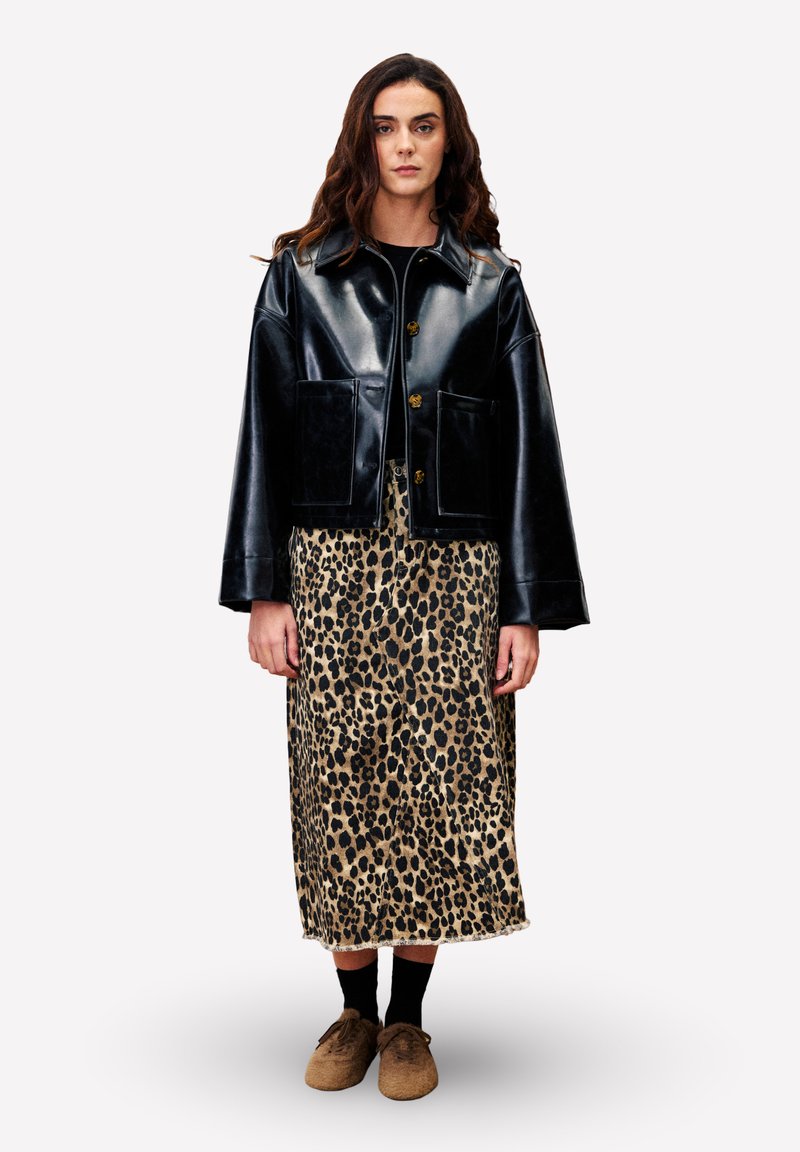 Vrouw staat gekleed in een zwart leren jack, een midirok met luipaardprint, zwarte sokken en een paarse schoenen tegen een effen witte achtergrond.