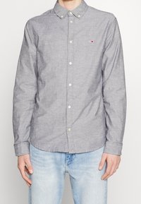 Gray button-down shirt with a tailored collar, long sleeves, and a small logo accent. Worn with light blue jeans. Smooth, woven texture.