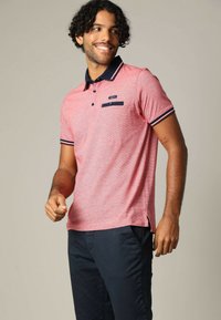 Rode, gestructureerde polo met een marineblauwe kraag en accenten, met een borstzak en zijsplitten. Gepaard met donkere broek.