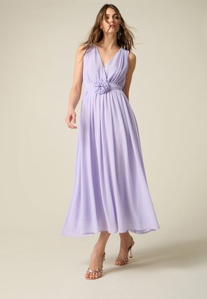 Frau trägt ein ärmelloses lavendelfarbenes Maxikleid mit Blumen-Gürtel-Detail, geht und posiert vor schlichtem neutralem Hintergrund.