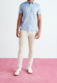 Light blue short-sleeve polo shirt, cream-colored skinny jeans, and white sneakers with black accents, against a pink carpeted background.
