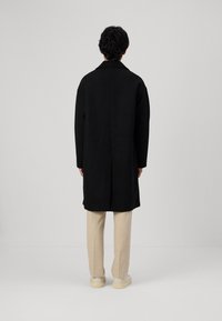 Schwarzer Wollmantel mit Reverskragen, geradem Schnitt und seitlichem Schlitz. Kombiniert mit beigen Hosen und hellen Sneakers.