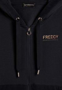 Felpa nera con zip realizzata in morbido materiale. Presenta un logo dorato, una cerniera metallica e cordini con punte in metallo. Mostrata distesa.