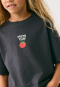 Černé bavlněné tričko s vyšitým designem. Text zní "ČAS NA PASTU" nad červenou rajčatou se zelenými listy. Krátké rukávy.