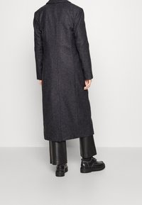 Abrigo largo de mezcla de lana en gris oscuro con corte recto, una abertura trasera y sin cierres visibles, combinado con pantalones de cuero negros y zapatos.