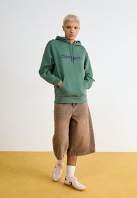 Grøn hætte sweatshirt med lilla logo, løse brune denim shorts og lyserøde sneakers. Sweateren har en lomme foran og snore.