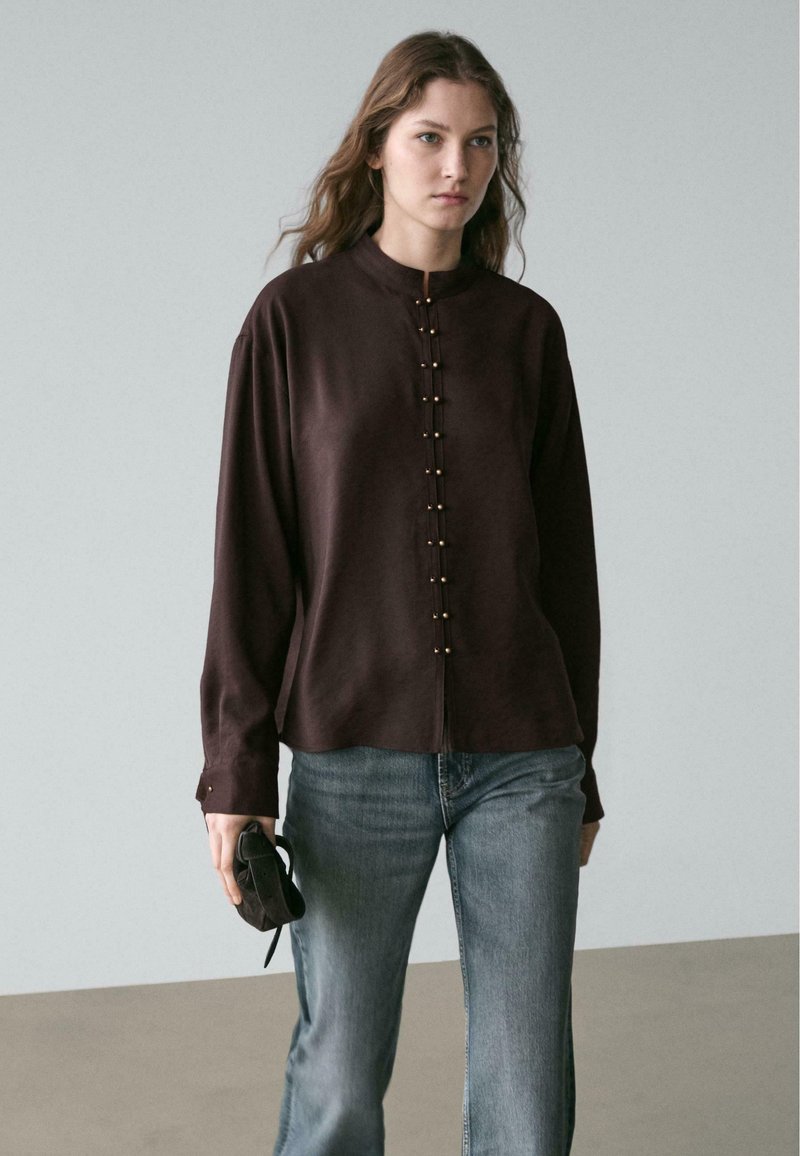 Dunkelbraune Bluse mit langen Ärmeln, einem Stehkragen und goldfarbenen Knopfdetails vorne, kombiniert mit hellblauen Denim-Jeans.