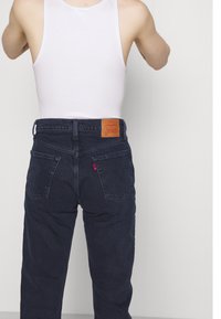 Vaqueros de mezclilla oscura con corte recto, que cuentan con dos bolsillos traseros, una etiqueta de cuero de marca y una pequeña etiqueta roja en el bolsillo trasero derecho.