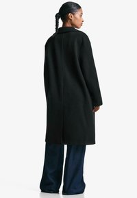 Zwarte lange jas met een losse pasvorm, voorzien van een kraag en zijzakken. Gladde textuur, eenvoudig ontwerp, gedragen over brede, donkere jeans.