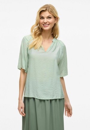 Blusa verde claro de cuello en V, fabricada en tejido texturizado, con mangas cortas y detalles fruncidos en los hombros, combinada con una falda a juego.