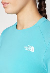 Chemise turquoise à manches courtes en tissu doux, dotée d'un logo blanc "The North Face" sur la poitrine gauche et d'un col rond.