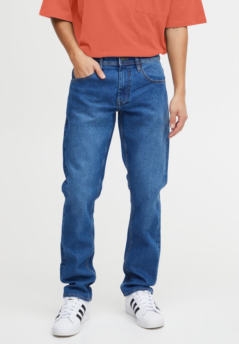 Vaqueros de mezclilla en azul medio con un diseño de pierna recta, cinco bolsillos y costuras en contraste. Combinados con zapatillas blancas con rayas negras.