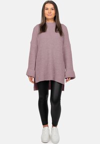 Vrouw staand met gezicht naar voren, gekleed in een oversized gebreide trui in mauve, zwarte leren legging en witte sneakers, tegen een witte achtergrond.
