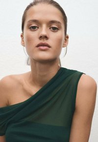 Dark green, sheer, one-shoulder top with draped fabric. Smooth texture, fitted shape, and minimalistic design against a neutral background.