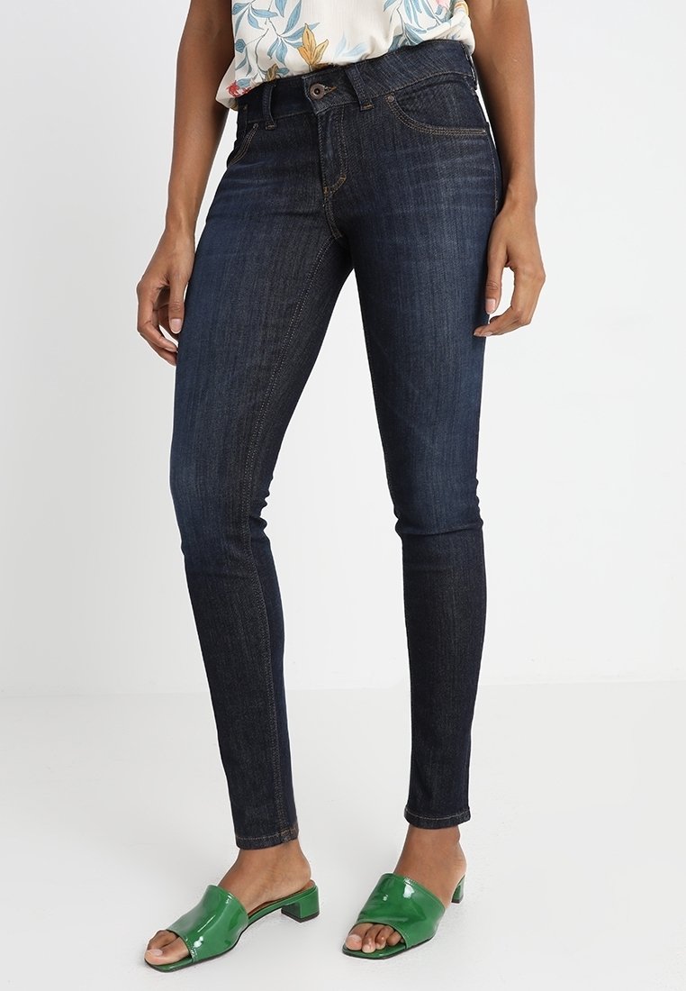 Skinny jeans in dark blue denim with orange stitching, featuring a standard five-pocket design and a fitted silhouette, paired with green mules.