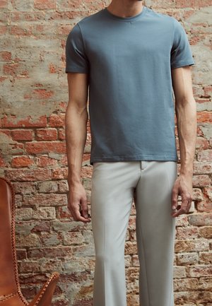 Homme portant un t-shirt ajusté à manches courtes bleu ardoise et un pantalon beige clair, debout devant un mur de briques usé.