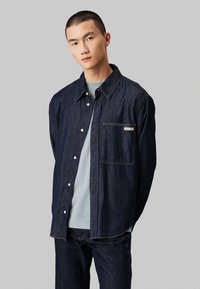 Camicia in denim di colore blu scuro con fronte a bottoni, colletto appuntito, taschino sul petto e cuciture in contrasto. Indossata sopra una T-shirt blu chiaro.