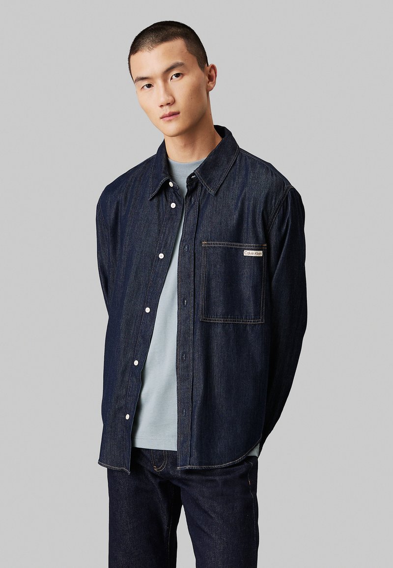 Camicia in denim di colore blu scuro con fronte a bottoni, colletto appuntito, taschino sul petto e cuciture in contrasto. Indossata sopra una T-shirt blu chiaro.