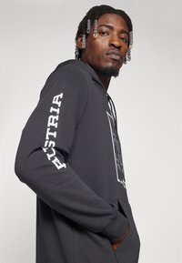 Graue Kapuzenjacke mit einem grafischen Design auf der Vorderseite, Kängurutasche und weißen Text auf dem Ärmel mit der Aufschrift "ÖSTERREICH". Aus weichem Stoff mit einer lockeren Passform.
