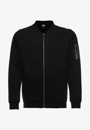 Veste bomber noire en tissu lisse, dotée d'une fermeture éclair à l'avant, de poignets côtelés et d'une poche zippée sur la manche gauche.