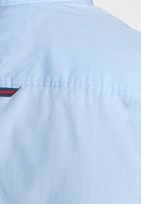 Ljust blå skjorttyg med en mjuk textur, med en liten röd och blå accentrand nära halsringningen och detaljerad sömnad.