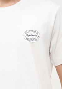 Άσπρο μπλουζάκι με μικρό ναυτικό μπλε τύπωμα λογότυπο στο αριστερό στήθος που γράφει «309 Portobello Rd Notting Hill Pepe Jeans Est. 1973.»