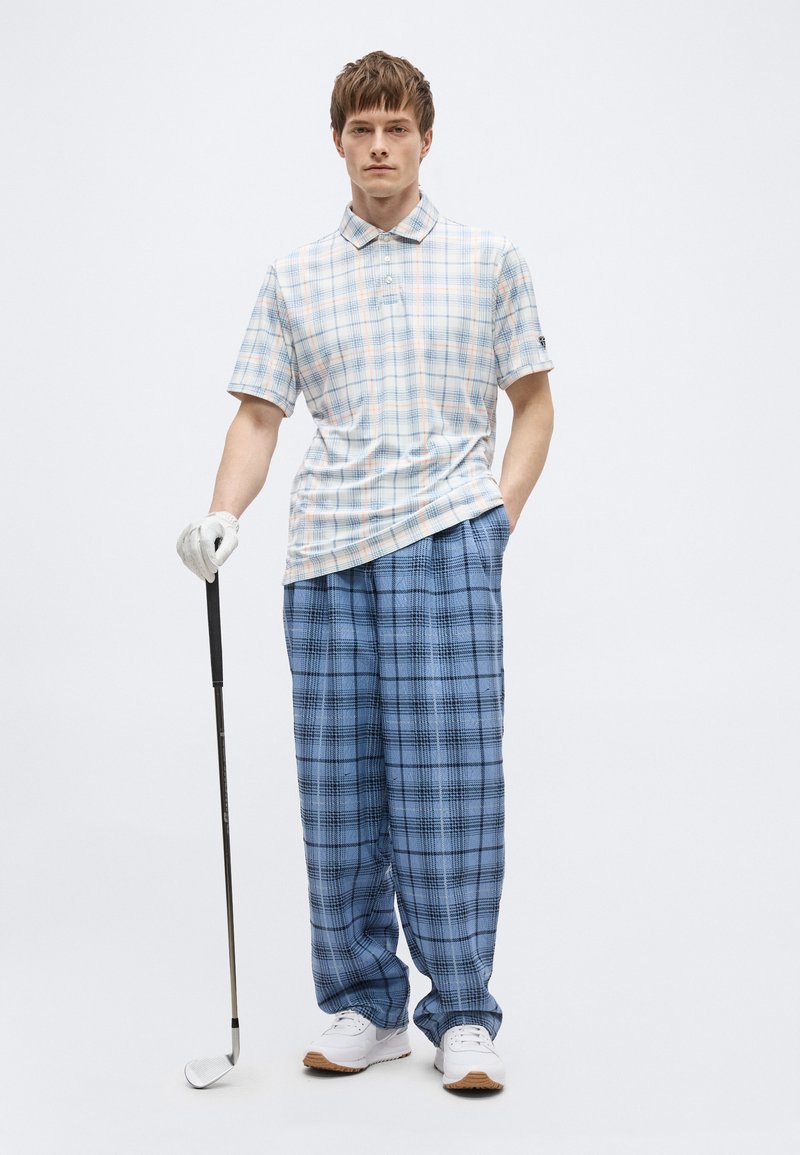 Hombre con polo blanco y a cuadros pastel y pantalones azules a cuadros, sosteniendo un palo de golf con un guante blanco en la mano izquierda.