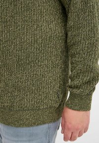Grüner Strickpullover mit strukturiertem Muster, kontrastierenden hellen Fäden, gerippten Bündchen und Saum sowie einem kleinen Logosticker an der Seite.