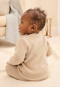 Pull en tricot beige à manches longues, avec un col rond et des coutures blanches, associé à un bas assorti. Enfant assis sur une surface texturée.
