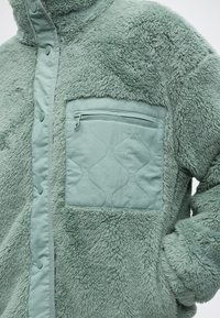 Μαλακό πράσινο fleece με φερμουάρ σε καπιτονέ τσέπη στο στήθος και κούμπωμα με κουμπιά μπροστά.