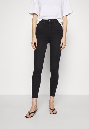 Woman wearing high-waist black skinny jeans, white loose t-shirt tucked in, and black heeled sandals standing against a plain light background.