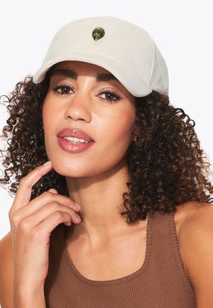 Femme aux cheveux bouclés portant une casquette beige avec un petit emblème métallique en forme de hibou et un haut marron sans manches à côtes, touchant son menton.