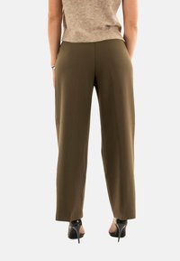 Donna che indossa pantaloni larghi verde oliva, un maglione senza maniche beige e scarpe con tacco nero, in piedi con le mani nelle tasche e voltata di spalle.
