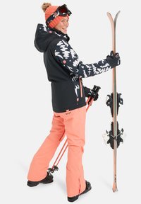 Conjunto de esqui com uma jaqueta preta com padrões brancos e rosas, calças laranja e um gorro de malha a condizer. Acessórios incluem bastões de esqui e esquís.