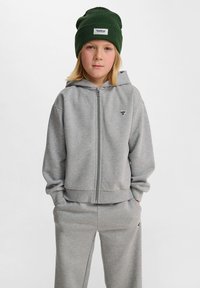 Grå zip-up hoodie och matchande sweatpants av mjukt material, med en liten logotyp på bröstet och ribbade manschetter vid handlederna och midjan.
