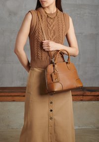 Borsa in pelle color tan con forma strutturata, manici superiori e tracolla rimovibile. Indossata con un top senza maniche in maglia e una lunga gonna con bottoni.