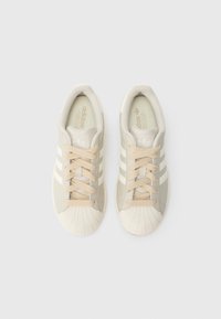 Tenisky s béžovo-bielou farebnou schémou, koženým zvrškom, okrúhlym gumovým zakončením na špičke a tromi bielymi pruhmi pozdĺž strán.