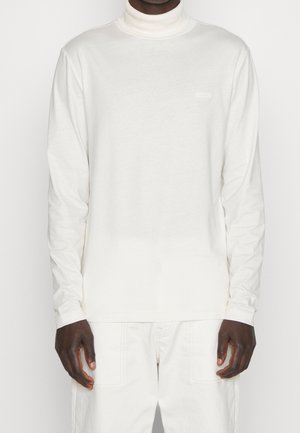 Mørkhudet person iført en hvid langærmet rullekravstrøje og hvide bukser, stående foran en ensfarvet baggrund.