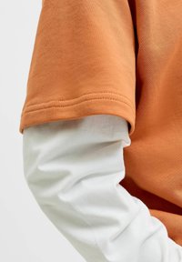 Vêtements superposés montrant un sous-vêtement blanc à manches longues sous une chemise orange à manches courtes, en se concentrant sur la zone du bras.