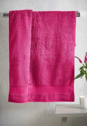 Lyserødt håndklæde foldet og hængende på en sølvfarvet håndklædeholder mod en lys væg, med et lille bord, der holder en hvid kop og en klar vase med pink blomster.