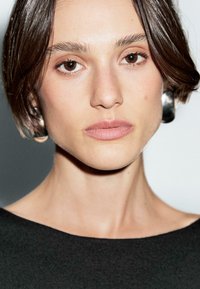 Giovane donna con capelli corti e scuri e trucco naturale, indossa una maglietta nera e grandi orecchini d'argento, fissa direttamente la fotocamera.