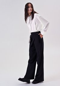 Blusa bianca a maniche lunghe con colletto, abbinata a pantaloni neri a gamba larga con fibbia. Scarpe platform nere completano il look.