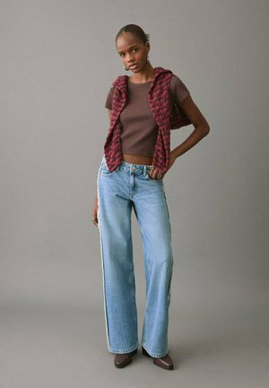 Crop top marrone a coste abbinato a un gilet fantasia rosso e nero, jeans larghi azzurri con spacchi laterali e scarpe marroni con punta a punta.