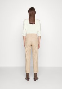 Beżowe dopasowane spodnie z wysokim stanem i prostymi nogawkami, w zestawieniu z kremową bluzką. Prezentują widok z tyłu z gładką teksturą i minimalnymi detalami.
