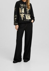 Sudadera negra con capucha que presenta gráficos dorados de Nirvana, combinada con pantalones anchos negros con rayas finas. El calzado es negro con un diseño sencillo.