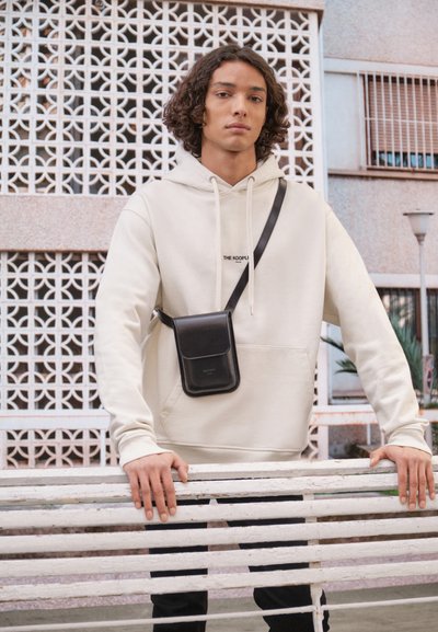 Haut à capuche beige clair avec une poche avant, sac bandoulière noir, debout sur un banc en bois blanc contre un mur à motifs.