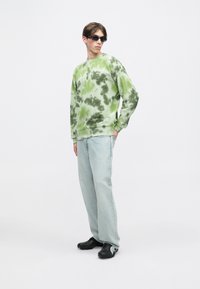 Πράσινη tie-dye μπλούζα με μακριά μανίκια, χαλαρή εφαρμογή; συνδυασμένη με απαλό μπλε τζιν και μαύρα αθλητικά παπούτσια. Μοντέλο που φορά μαύρα γυαλιά ηλίου.