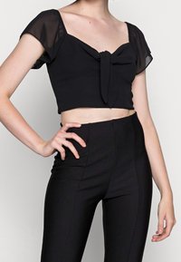 Svart avkortad topp med genomskinliga korta ärmar, med detaljer i form av en rosett framtill, tillsammans med högmidjade svarta tajta byxor. Slät tygtextur.